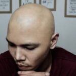 Bald man sitting indoors, hands together, with motivational quotes on the wall.