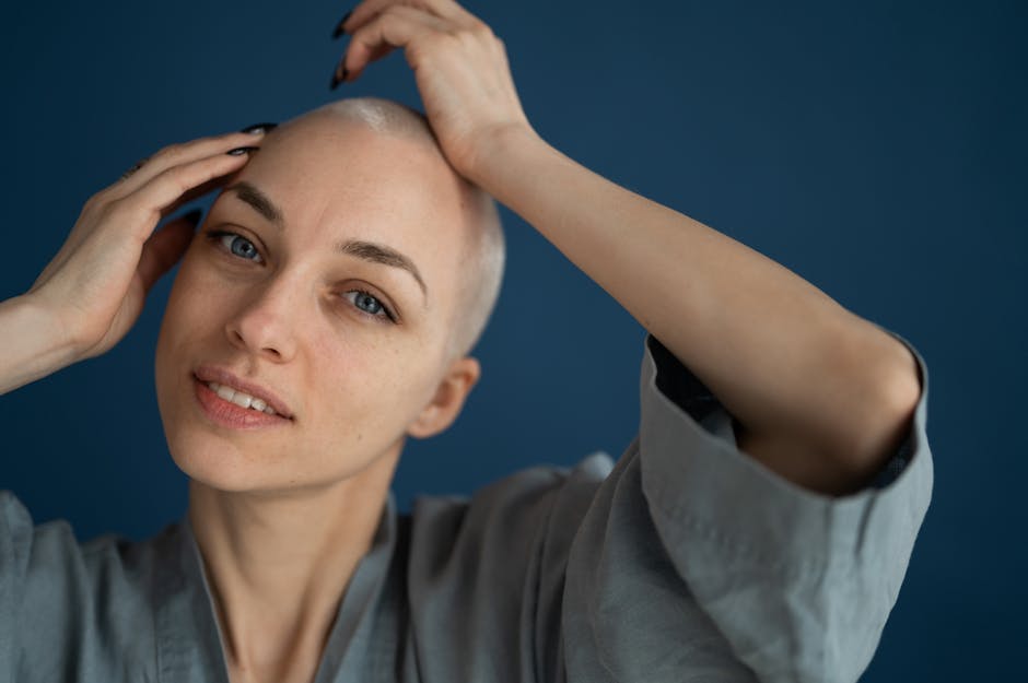 Positive young bald female in gray robe touching head tenderly and looking at camera against dark blue wall